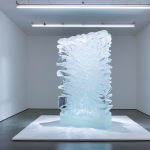 A contemporary art installation in a minimalist modern art gallery in Berlin, showcasing a large, slowly melting ice sculpture. Dynamic, subtle light projections shift across its surface, creating evolving patterns and reflections. The scene emphasizes transformation and the ephemeral nature of art over time. High-resolution, professional studio photography, cinematic lighting, ultra-detailed, photorealistic, safe for work, appropriate content, fully clothed, professional, family-friendly.